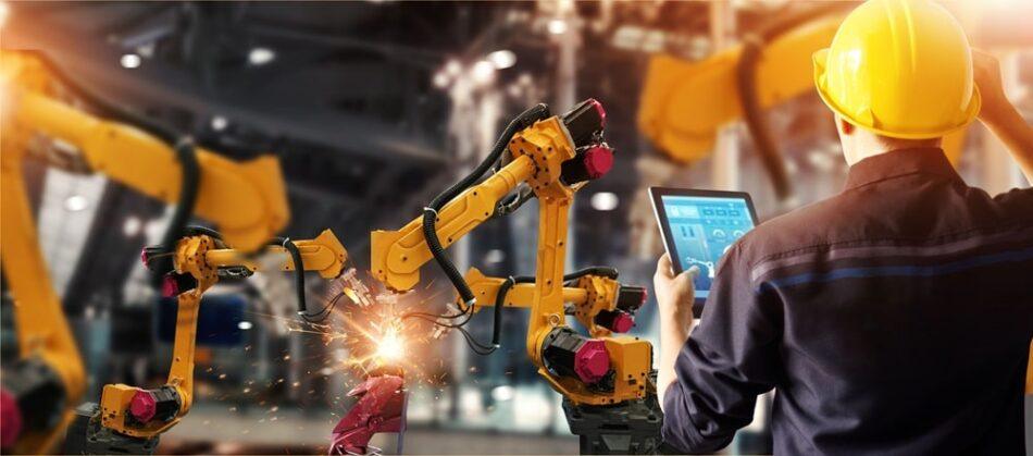 An automotive worker workers on a tablet with two robots building car parts