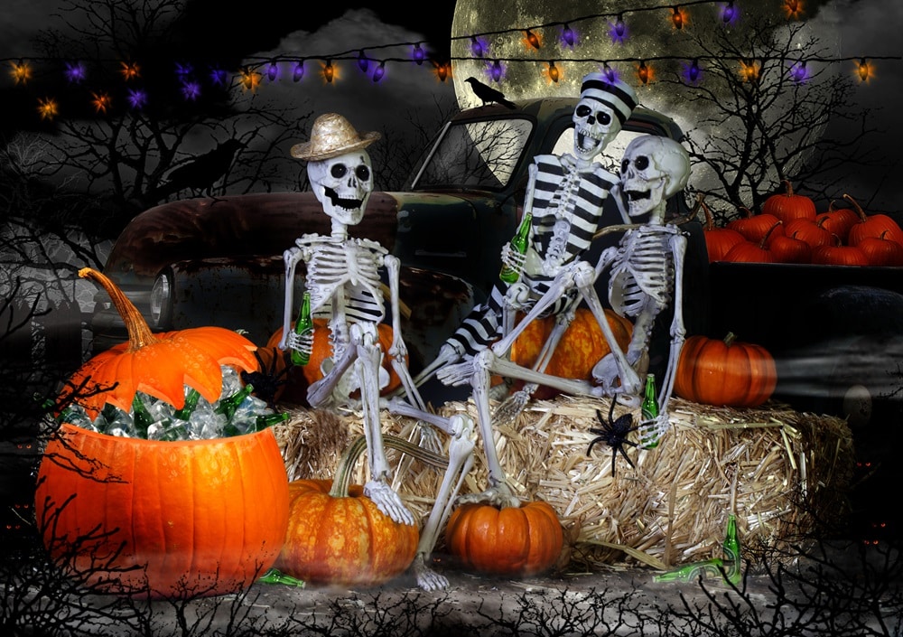3 spooky skeletons sat on a bench with lots of pumpkins and haloween lights, and a full moon in the back at night with a black sky