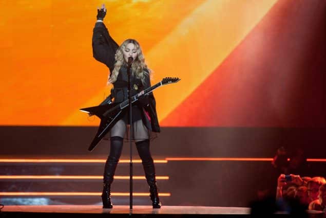 Madonna onstage with an orange backdrop, she's got a black flying V guitar