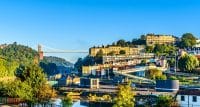View of Clifton Suspension Bridge spanning the Avon Gorge with Bristol's scenic cityscape and lush greenery under a clear blue sky